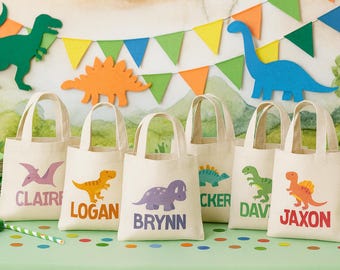 Puede incluir: Seis bolsas de tela beige con asas, cada una con un gráfico de dinosaurio de colores y un nombre impreso. Los nombres Claire, Logan, Brynn, Cker, Dav y Jaxon aparecen en varios colores y fuentes en la parte frontal de cada bolsa.