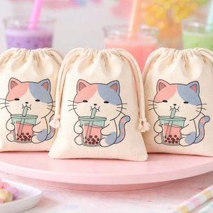 May include: Three beige drawstring bags featuring a cartoon cat design. The cat is pink and blue, holding a boba tea. The bags are on a pink cake stand, with cupcakes and drinks in the background.