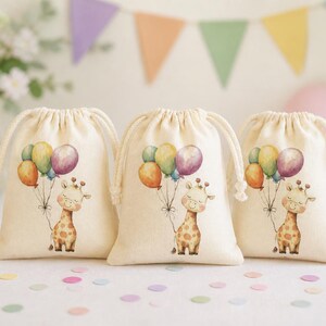 Może przedstawiać: Trzy beżowe worki ze sznurkiem z ilustracją żyrafy trzymającej kolorowe balony. Balony są żółte, pomarańczowe, fioletowe i zielone. Worki znajdują się na białej powierzchni z konfetti i dekoracjami imprezowymi w tle.