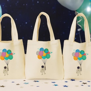 Può includere: Tre borse tote in tela color crema con un'illustrazione colorata di un astronauta che fluttua con palloncini a forma di pianeta. Le borse hanno una forma rettangolare con manici. Lo sfondo è a tema spaziale.
