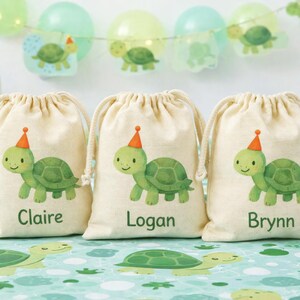 Op de afbeelding: Drie beige trekkoordzakken met een groene schildpadillustratie met een feesthoed. Elke tas heeft een andere naam: Claire, Logan en Brynn. De achtergrond bevat ballonnen en een taart.