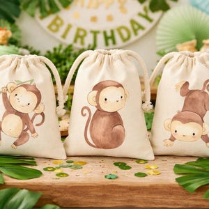 May include: Three beige drawstring bags with adorable cartoon monkey illustrations. Each bag features a different monkey pose in shades of brown. The bags are part of a jungle-themed birthday party setup.
