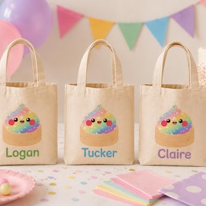 May include: Three beige canvas tote bags with two handles each. Each bag features a colorful, smiling rainbow-colored cupcake graphic with stars. The bags are personalized with the names Logan in green, Tucker in blue, and Claire in purple.