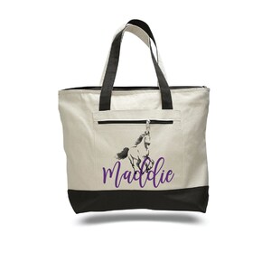 Pode incluir: Uma bolsa tote de lona branca com al&ccedil;as pretas e fundo preto. A bolsa tem um bolso com z&iacute;per na frente e um gr&aacute;fico de cavalo roxo com o nome "Maddie" impresso abaixo.