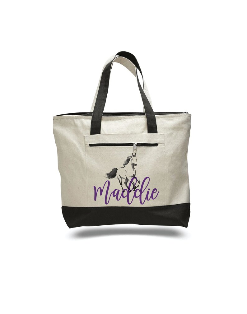 Pode incluir: Uma bolsa tote de lona branca com al&ccedil;as pretas e fundo preto. A bolsa tem um bolso com z&iacute;per na frente e um gr&aacute;fico de cavalo roxo com o nome "Maddie" impresso abaixo.