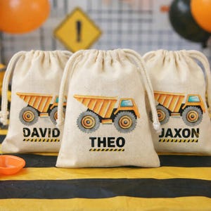 May include: Three beige drawstring bags with a yellow dump truck design and personalized names: David, Theo, and Jaxon. The bags are on a yellow and black striped surface, with construction-themed decorations in the background.