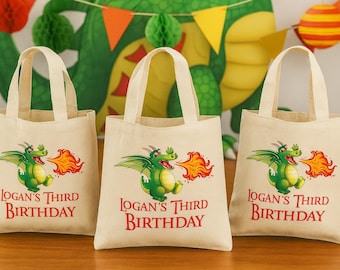 May include: Three beige canvas tote bags with handles, each featuring a green dragon breathing fire. The text "Logan's Third Birthday" is printed in red below the dragon illustration. Party decorations are in the background.
