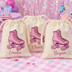 Puede incluir: Tres bolsas beige con cordón con ilustraciones de patines rosas y nombres: Emalee, Claire y Brynn. Las bolsas están sobre una superficie a cuadros rosa y blanco, con decoraciones de fiesta en el fondo.