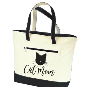 May include: A cream-colored canvas tote bag with black handles and a black bottom. The bag has a black cat face with whiskers and the text "Cat Mom" printed on the front.