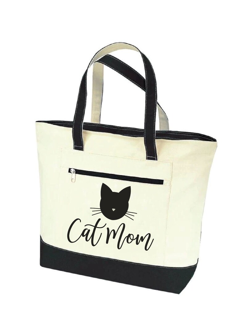 May include: A cream-colored canvas tote bag with black handles and a black bottom. The bag has a black cat face with whiskers and the text "Cat Mom" printed on the front.