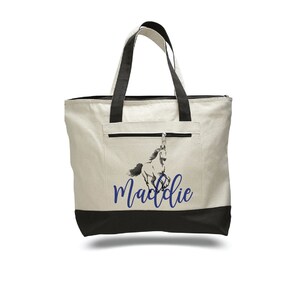 Pode incluir: Uma bolsa tote de lona branca com al&ccedil;as pretas e fundo preto. A bolsa tem um bolso com z&iacute;per na frente e um gr&aacute;fico azul de um cavalo com o nome "Maddie" impresso abaixo.