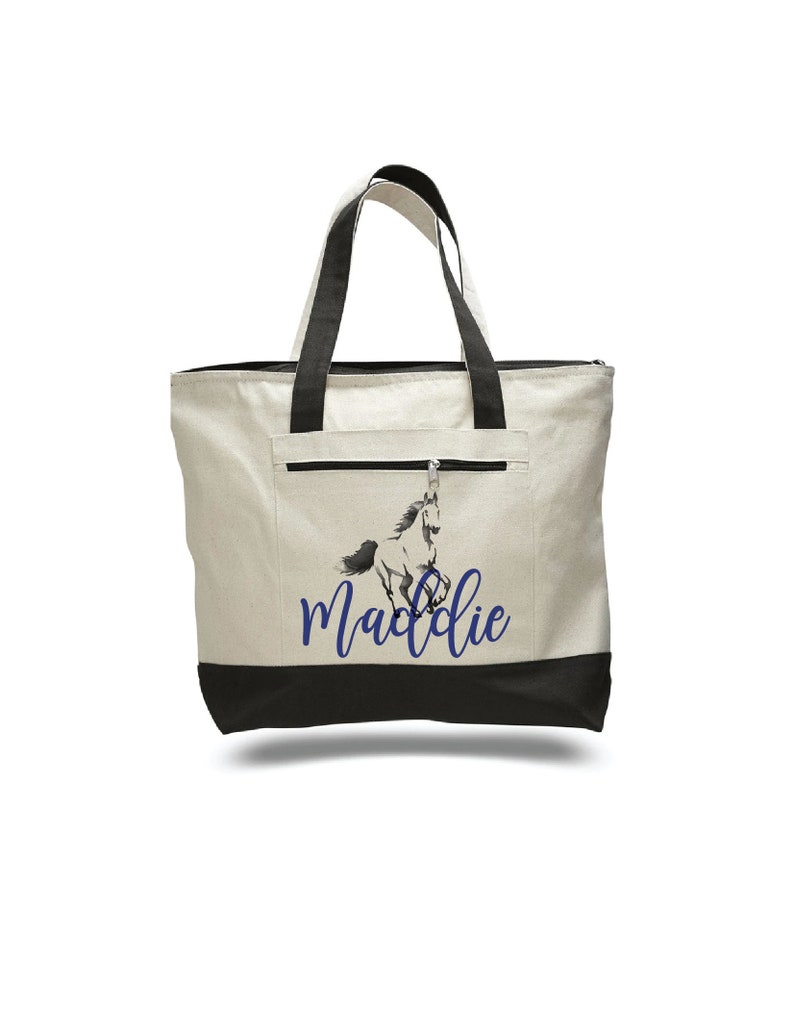 Pode incluir: Uma bolsa tote de lona branca com al&ccedil;as pretas e fundo preto. A bolsa tem um bolso com z&iacute;per na frente e um gr&aacute;fico azul de um cavalo com o nome "Maddie" impresso abaixo.