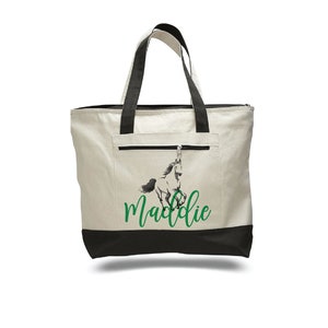 Pode incluir: Uma bolsa tote de lona branca com al&ccedil;as pretas e fundo preto. A bolsa tem um bolso com z&iacute;per na frente e apresenta um gr&aacute;fico de cavalo preto e branco com o nome "Maddie" em texto verde.