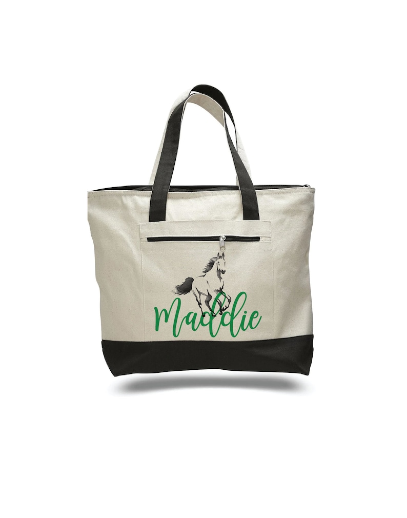 Pode incluir: Uma bolsa tote de lona branca com al&ccedil;as pretas e fundo preto. A bolsa tem um bolso com z&iacute;per na frente e apresenta um gr&aacute;fico de cavalo preto e branco com o nome "Maddie" em texto verde.