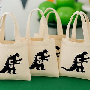 Può includere: Diverse borse tote in tela beige con manici coordinati. Ogni borsa presenta una silhouette nera di un mostro simile a Godzilla con il numero 5. Le borse sono disposte su una superficie verde, suggerendo un tema di festa di compleanno.