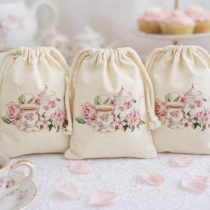 May include: Three cream-colored drawstring bags with a watercolor design of a teapot, teacup, and pink roses. The bags are arranged on a white surface with scattered pink rose petals. A teacup and saucer are in the foreground.