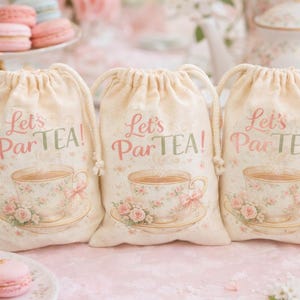 May include: Three cream-colored drawstring bags with the text "Let's ParTEA!" in pink and green. Each bag features a floral teacup design. The background includes pastries and teacups, suggesting a tea party theme.