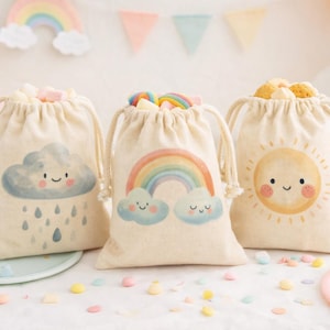 Puede incluir: Tres bolsas beige con cordón, con ilustraciones de una nube, un arcoíris y un sol. Cada bolsa está llena de golosinas y se encuentra sobre un plato pastel. El fondo incluye cupcakes, globos y una pancarta de arcoíris.
