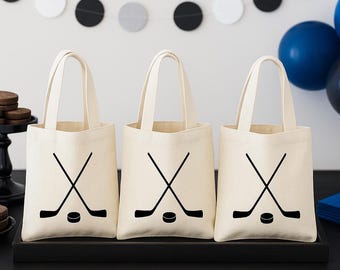Puede incluir: Tres bolsas de mano color crema con gráficos de palos y discos de hockey negros. Las bolsas tienen asas resistentes y están dispuestas sobre una bandeja negra. El fondo incluye una guirnalda de círculos negros, blancos y grises, y globos azules.