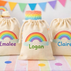 Könnte beinhalten: Drei beigefarbene Kordelzugbeutel mit Regenbogen-Design und personalisierten Namen: Emalee, Logan und Claire. Die Beutel haben einen bunten Regenbogen über einer Wolke. Im Hintergrund sind ein Regenbogenkuchen und Partydekorationen zu sehen.