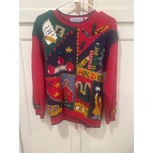 Könnte beinhalten: Ein farbenfroher Pullover zum Thema Schule mit roten Ärmeln und einer gemusterten Vorderseite. Das Design umfasst Bücher, Äpfel, das ABC und ein Schulhaus. Der Pullover hat einen Rundhalsausschnitt und hängt an einem weißen Kleiderbügel.