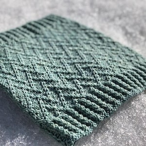 May include: A square, knitted item in a muted teal color. The texture features a repeating diamond pattern in the center, with a ribbed border. The item is resting on a textured, white surface, possibly snow or ice.