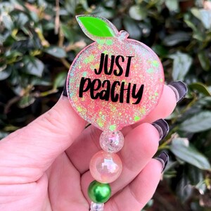 May include: A pink peach-shaped badge holder with glitter and the text "Just Peachy". The holder has a green leaf and is attached to a clear retractable badge reel with three beads: a clear bead, a pink iridescent bead, and a green bead.
