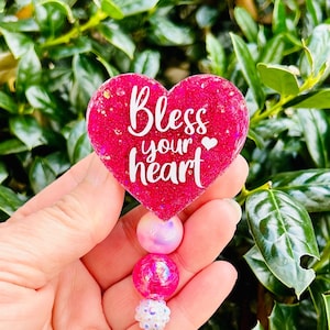 May include: Pink heart-shaped resin charm with glitter and the text "Bless your heart" with a small heart icon. The charm is attached to a clear badge holder with a pink, white, and iridescent bead chain.