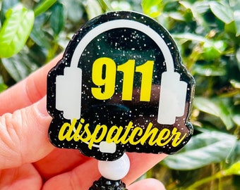 以下が含まれることがあります： 黒と黄色の911ディスパッチャーバッジリール、黒と白のビーズチェーン付き。バッジリールは黒の背景に白いヘッドホン、黄色の「911ディスパッチャー」の文字が書かれています。