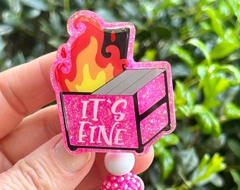 May include: Pink glitter badge reel with a pink and black image of a burning matchbox with the text "IT'S FINE". The reel has pink, white, and clear beads.