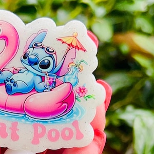 May include: A white and pink glitter badge reel with a cartoon image of Stitch from Lilo & Stitch on a pink flamingo float. The text "Float Pool" is printed on the badge reel. The badge reel has a white, blue and white swirl, and pink glitter beads.