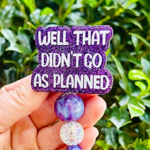 Peut inclure: Badge en paillettes violettes avec le texte "WELL THAT DIDN'T GO AS PLANNED" attaché à un porte-badge rétractable à perles violettes, blanches et transparentes.