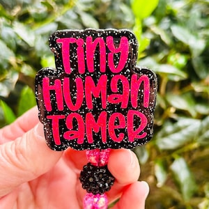 May include: Black and pink glitter badge holder with the text "Tiny Human Tamer" in pink lettering. The badge holder has a black and pink beaded chain with a clear plastic clip.