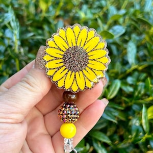 Puede incluir: Un portatarjetas de girasol con un diseño de resina brillante en amarillo y marrón. El cordón retráctil está decorado con tres cuentas: una marrón sólida, una marrón con incrustaciones de diamantes de imitación y una amarilla sólida.