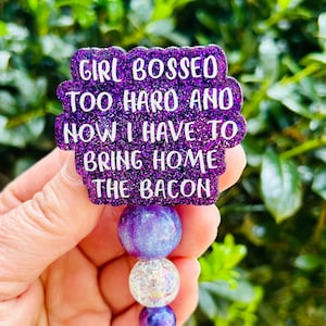 May include: Purple glitter badge reel with a white text overlay that reads "Girl Bossed Too Hard And Now I Have To Bring Home The Bacon". The reel has a purple, clear, and iridescent bead charm.