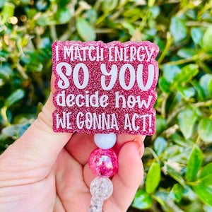 Puede incluir: Un portatarjetas cuadrado con fondo magenta brillante y texto blanco que dice I MATCH ENERGY SO YOU DECIDE HOW WE GONNA ACT! El portatarjetas cuenta con un cordón retráctil unido a una cuenta blanca y una cadena de cuentas rosas y transparentes.