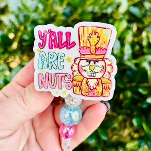 May include: A badge reel with a white background featuring a cartoon nutcracker in a pink and gold uniform. The text "Y'ALL ARE NUTS" is written in pink and blue. The reel has a beaded design with pink, blue, and silver beads.