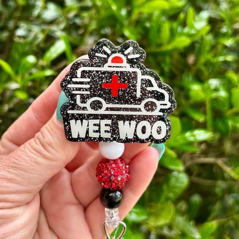 May include: A black and silver glitter badge reel featuring an ambulance design with a red cross and the words "WEE WOO" in white. The badge reel has a red, white, and black beaded accent.
