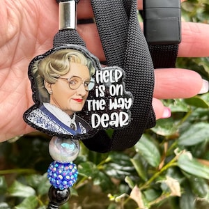 Puede incluir: Lanyard negro con un portagafete transparente y un dije con la cara de una mujer con el texto "Help is on the way dear".