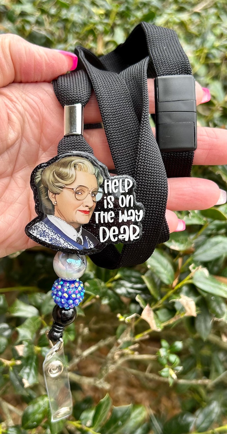 Puede incluir: Lanyard negro con un portagafete transparente y un dije con la cara de una mujer con el texto "Help is on the way dear".