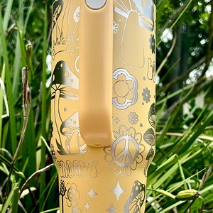 May include: A yellow stainless steel travel mug with a white lid and a yellow handle. The mug features a groovy 70s design with mushrooms, peace signs, and flowers. The text "Groovy" is printed on the mug.