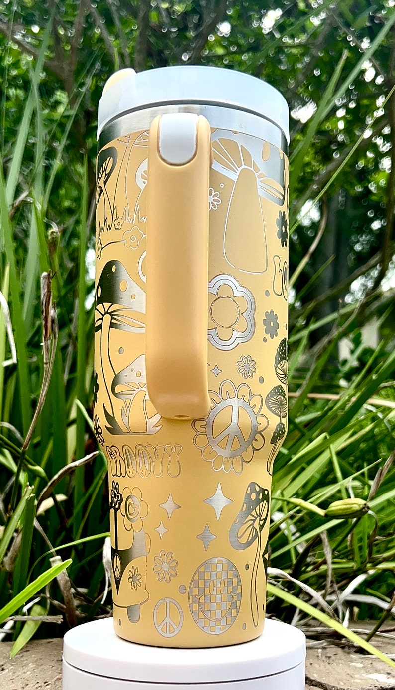 May include: A yellow stainless steel travel mug with a white lid and a yellow handle. The mug features a groovy 70s design with mushrooms, peace signs, and flowers. The text "Groovy" is printed on the mug.