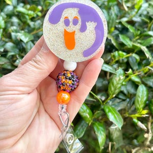 Puede incluir: Un portacredenciales circular con fondo blanco brillante y un gr&aacute;fico de fantasma morado con ojos y boca naranjas. El carrete est&aacute; unido a una cadena de cuentas con una cuenta blanca, una cuenta de diamantes de imitaci&oacute;n multicolor y una naranja.