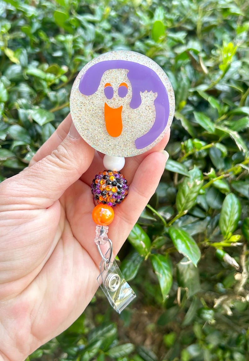 Puede incluir: Un portacredenciales circular con fondo blanco brillante y un gr&aacute;fico de fantasma morado con ojos y boca naranjas. El carrete est&aacute; unido a una cadena de cuentas con una cuenta blanca, una cuenta de diamantes de imitaci&oacute;n multicolor y una naranja.