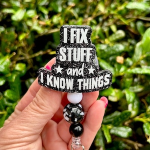 May include: A black glittery badge reel featuring the white text I FIX STUFF and I KNOW THINGS with two white stars. The badge reel is attached to a beaded lanyard component consisting of one white bead, one black and white patterned bead.