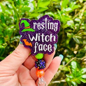 May include: A badge reel featuring a purple glitter acrylic topper with the white text "resting witch face," a green witch hat, and an orange bat. Below the topper is a vertical strand of three beads: one lime green, one black with purple and orange rhinestones, and one bright orange.
