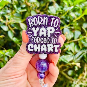 May include: A purple glitter badge reel with the text "BORN TO YAP FORCED TO CHART" in white. The badge reel has a white bead, a large purple bead, and a smaller purple bead.