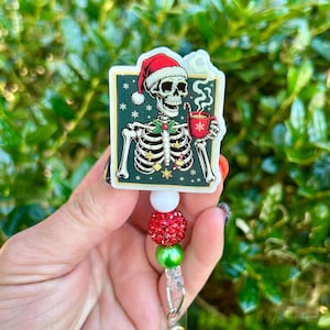 May include: A rectangular badge reel featuring a skeleton wearing a red and white Santa hat, holding a red mug with a candy cane. The background is dark green with white snowflakes. Below the image are three beads: one white, one red, and one green.