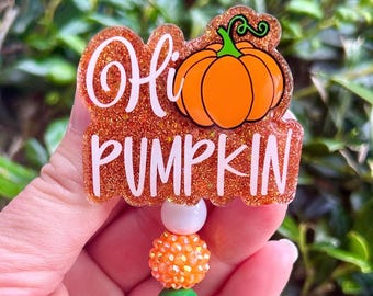 May include: A badge reel featuring an orange glitter acrylic topper with the white text Hi PUMPKIN and a graphic of an orange pumpkin. Below the topper are three beads in white, orange, and green, attached to a retractable cord mechanism.