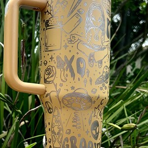May include: A beige travel mug with a handle, featuring etched illustrations of aliens, mushrooms, a peace sign, and the word "GROOVY." The mug has a white lid and a stainless steel rim.